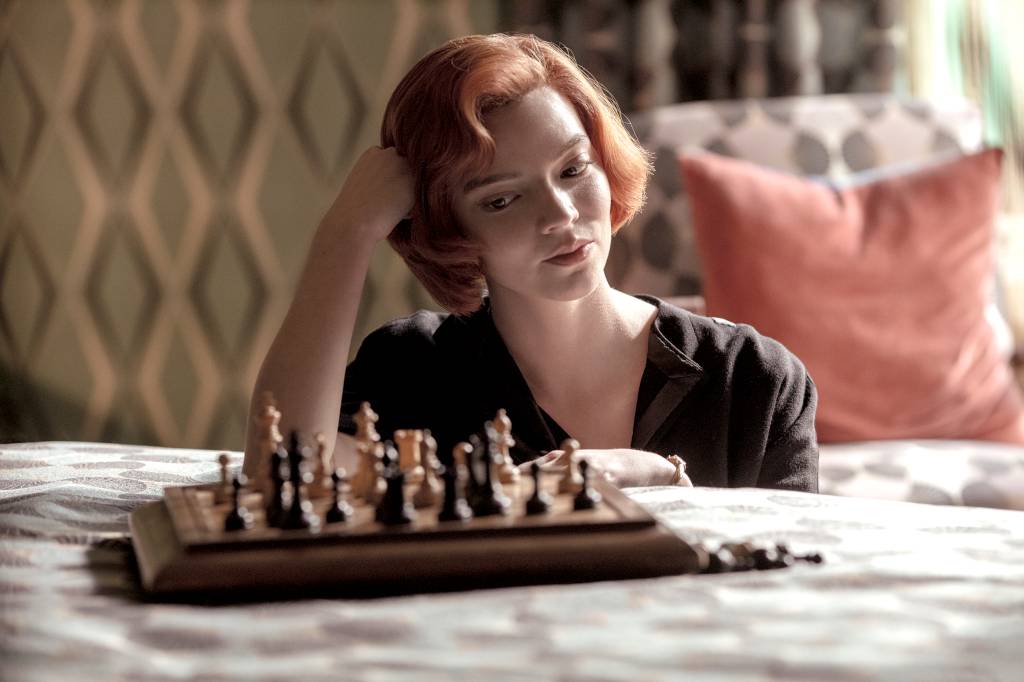 Atriz que interpreta Beth Harmon olha para um tabuleiro de xadrez em cima da cama e sorri com a mão apoiada na cabeça