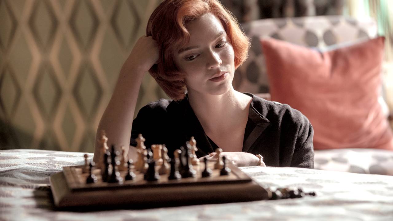 Atriz que interpreta Beth Harmon olha para um tabuleiro de xadrez em cima da cama e sorri com a mão apoiada na cabeça