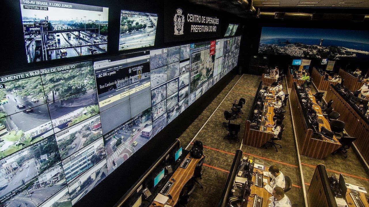 Tela como de cinema mostra imagens de câmeras do Rio de Janeiro, na frente pessoas estão sentadas em fileiras de mesas e utilizando o computador
