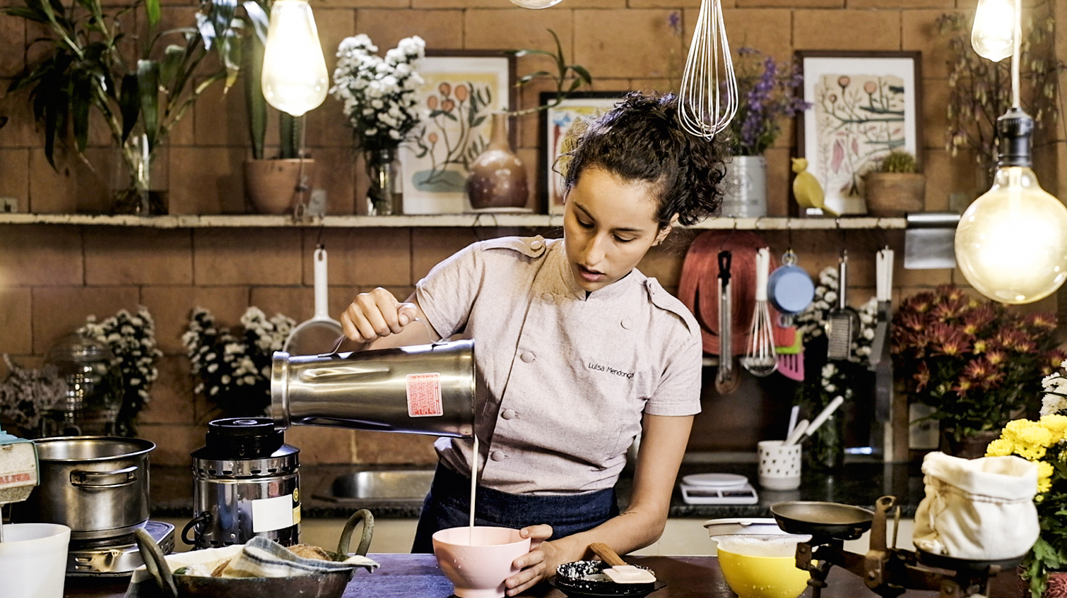 A imagem mostra a confeiteira vegana Luisa Mendonça na cozinha, despejando numa vasilha o leite vegetal que está no copo de um liquidificador