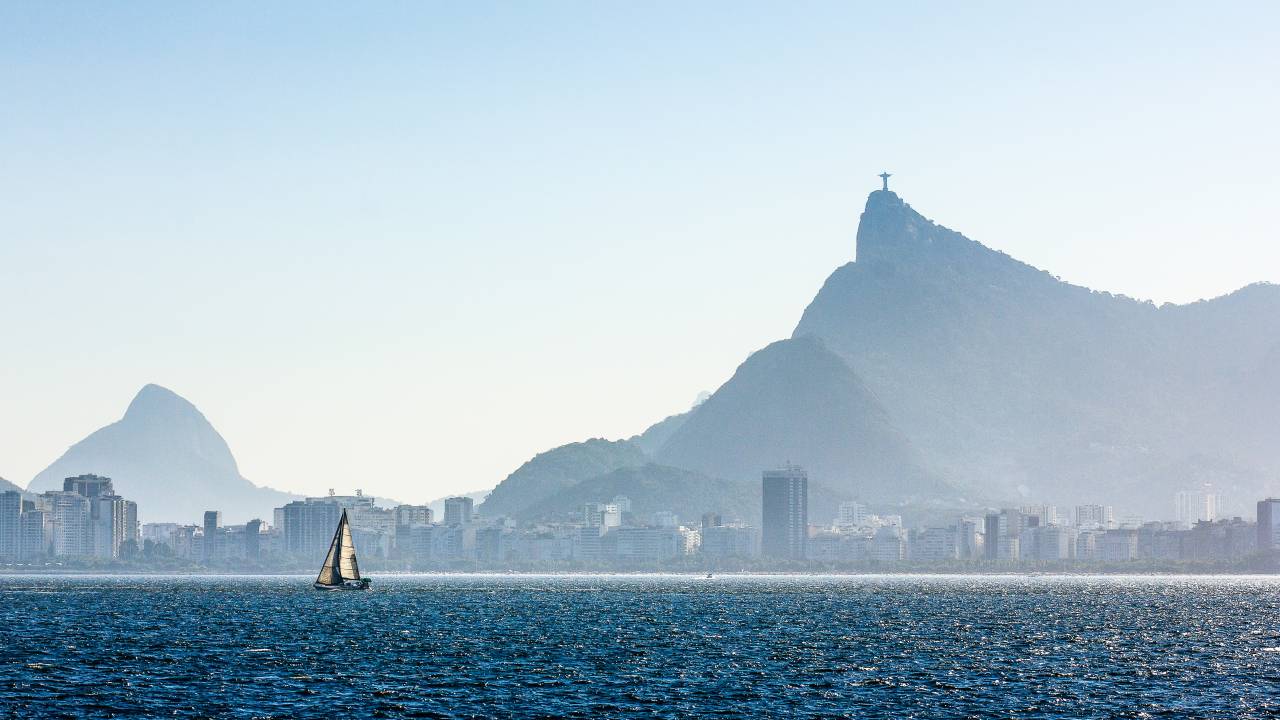 A imagem, em tons de azul, mostra um barco velejando com o relevo do Rio e o Cristo Redentor ao fundo