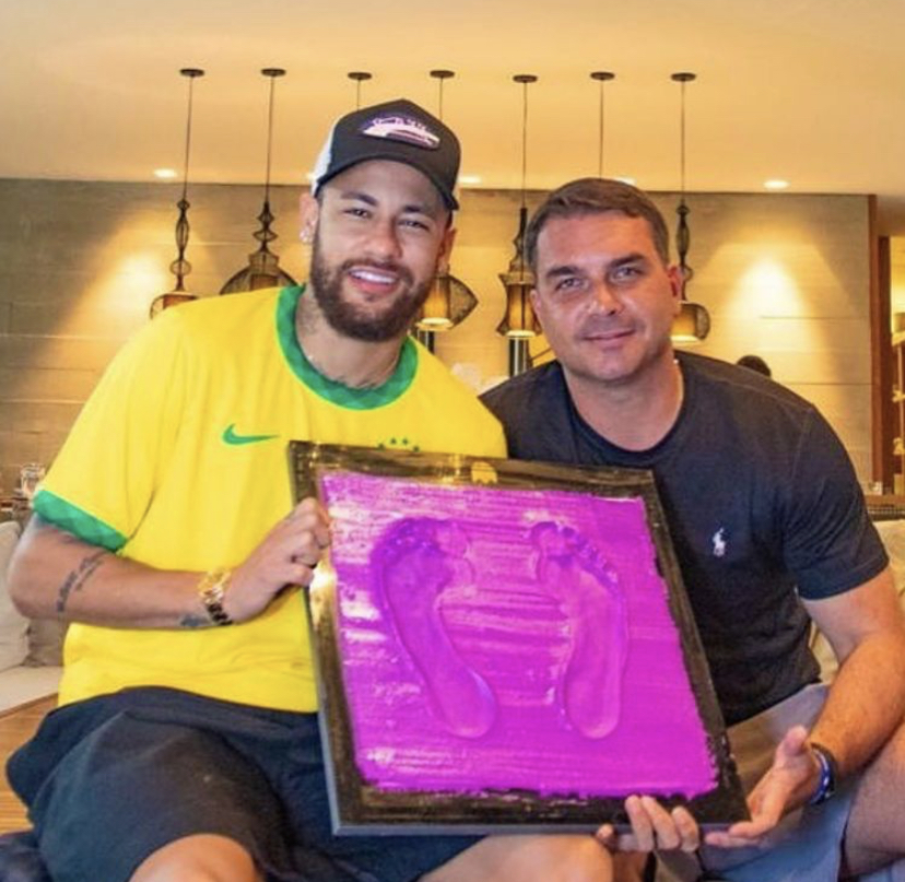 A imagem mostra o jogador de futebol Neymar sentado ao lado de Flávio Bolsonaro; ambos seguram o molde com a marca de seus pés, que entraram na calçada da fama do Maracanã
