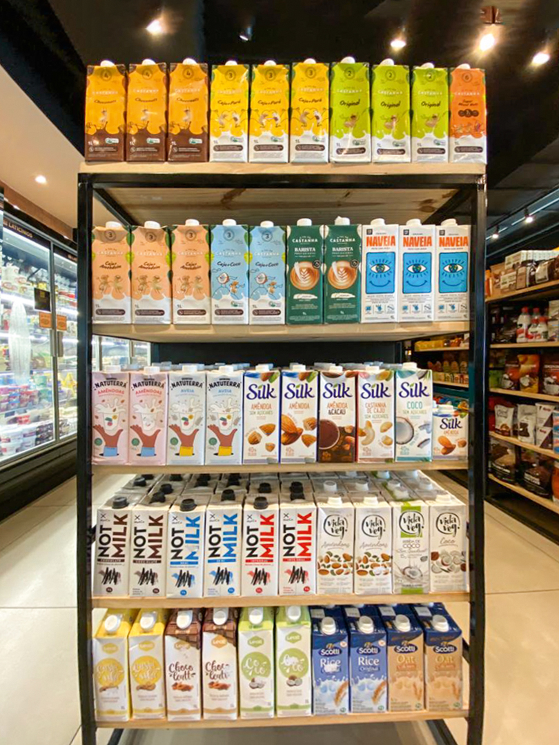 A imagem mostra uma seção de leites vegetais de um mercado com várias marcas disponíveis nas prateleiras