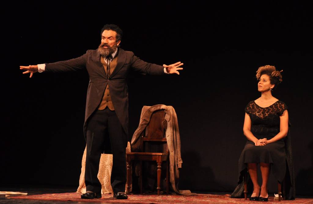 Ator Deo Garcez no palco com os braços abertos e roupas de época. Ao fundo, atriz está sentada
