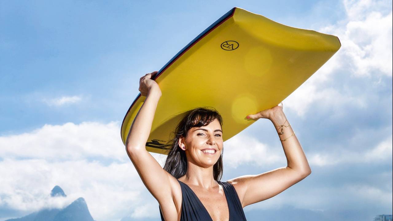 A imagem mostra a jornalista e campeã mundial de bodyboarding Gelnda Kozlowski sorrindo na praia, num dia de sol, com a prancha apoiada na cabela