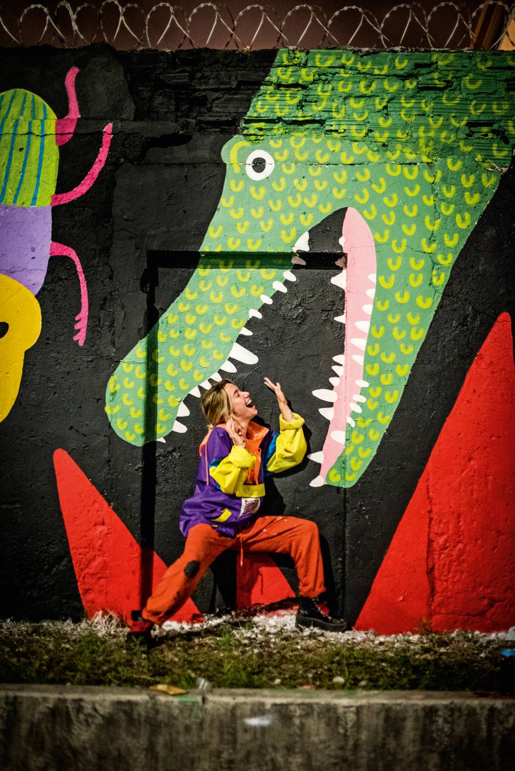A imagem mostra a artista Chica Capeto interagindo com um mural pintando por ela, como es estivesse dentro da boca de um jacaré que pintou na Zona Portuária