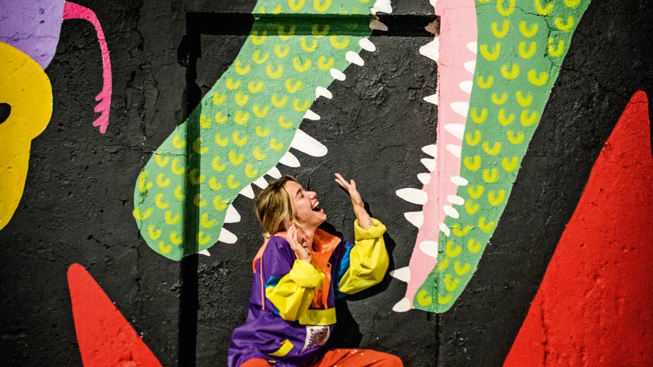 A imagem mostra a artista Chica Capeto interagindo com um mural pintando por ela, como es estivesse dentro da boca de um jacaré que pintou na Zona Portuária