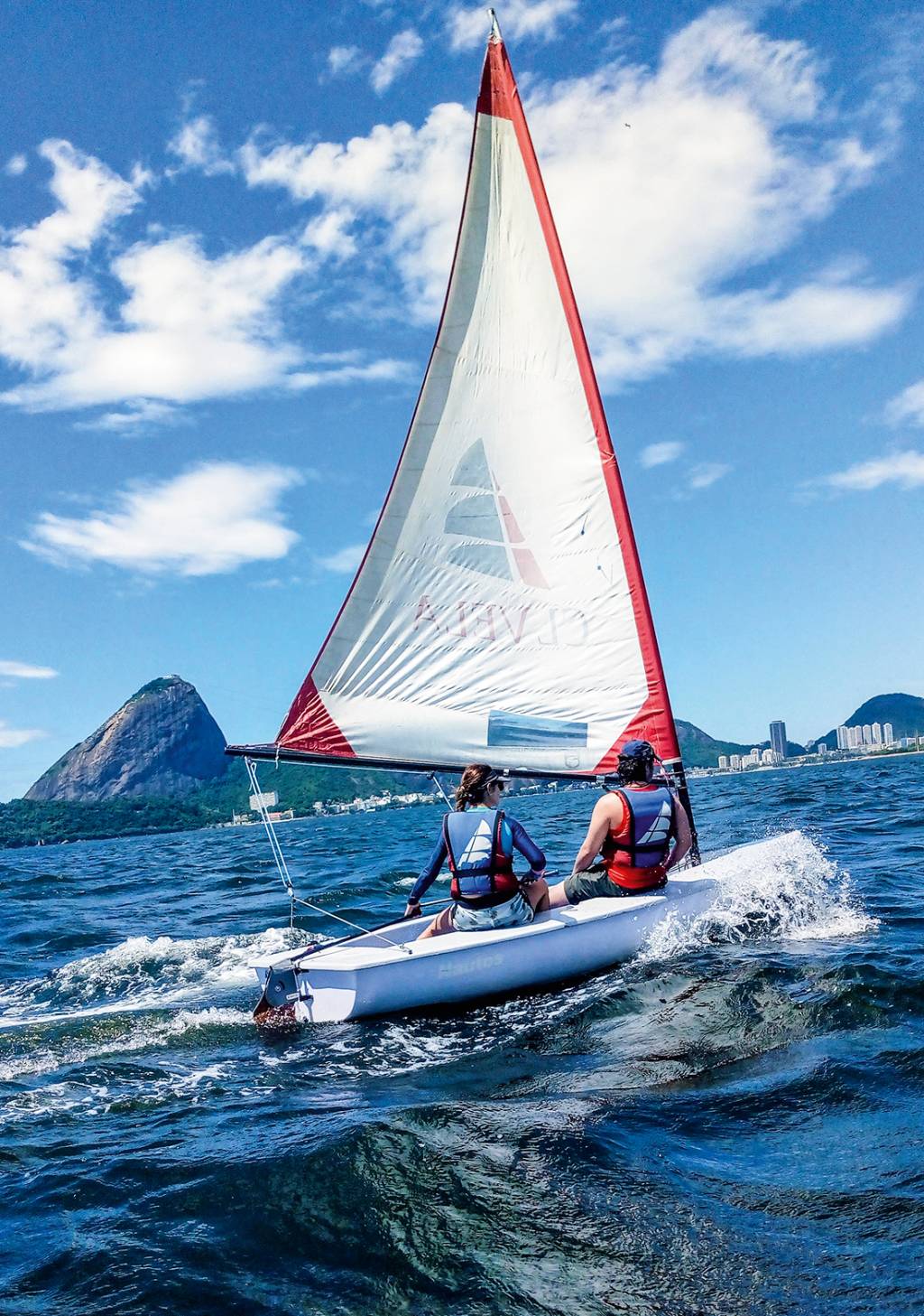 A imagem mostra duas pessoa num dingue, barco a vela, nas águas da Baía de Guanabara