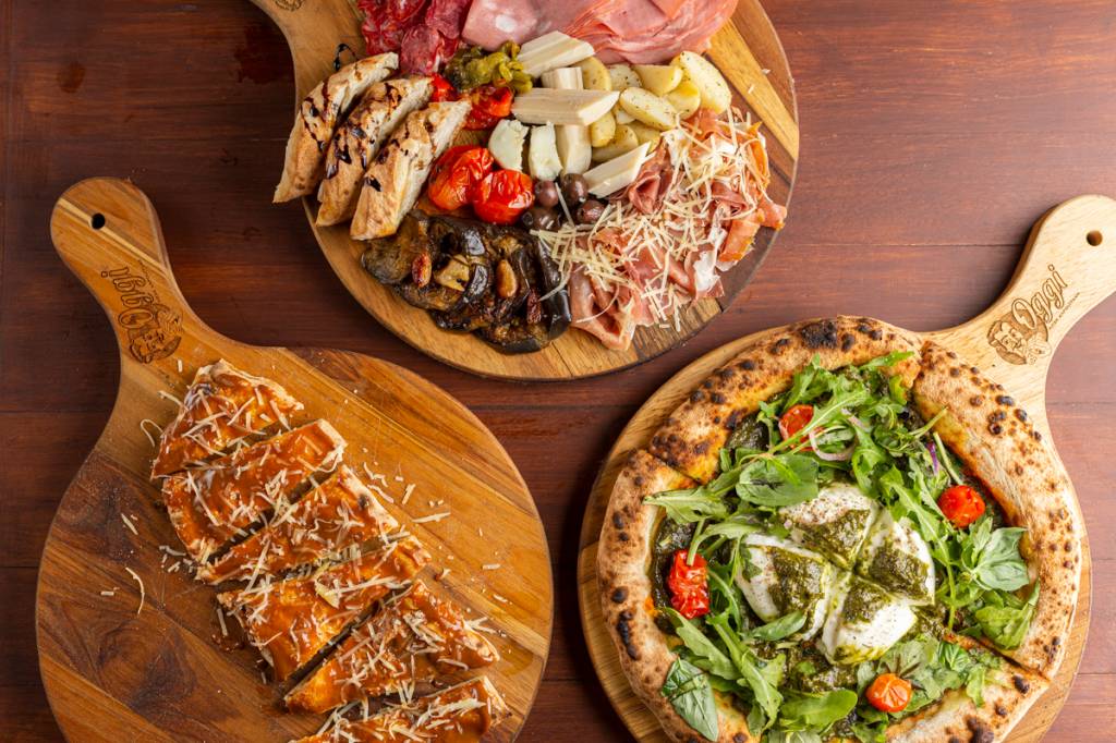 A imagem mostra três tábuas com pizza, frios e massa de pão