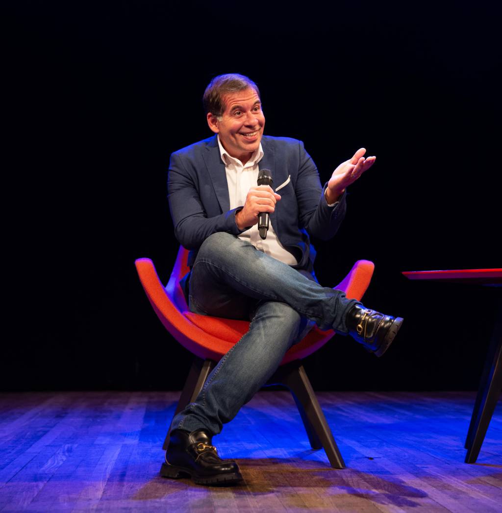 Leandro Hassum, no palco, sentado numa cadeira, de pernas cruzadas, com microfone em punho
