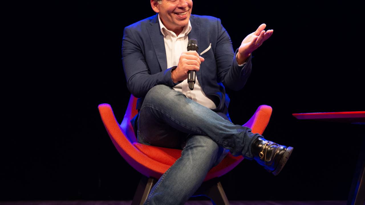 Leandro Hassum, no palco, sentado numa cadeira, de pernas cruzadas, com microfone em punho