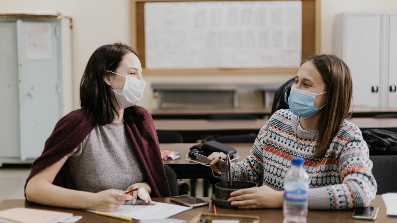 A imagem mostra duas alunas de máscara sentadas numa sala de aula