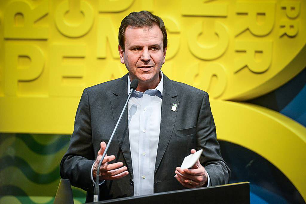 A imagem mostra o prefeito Eduardo Paes falando ao microfone numa bancada com fundo amarelo