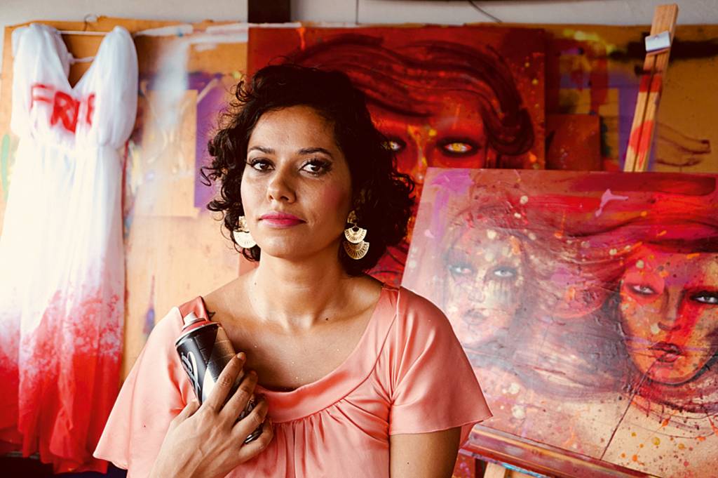 A imagem mostra a artista Panmela Catsri à frente de um de seus trabalhos, com uma lata de tinta em spray na mão
