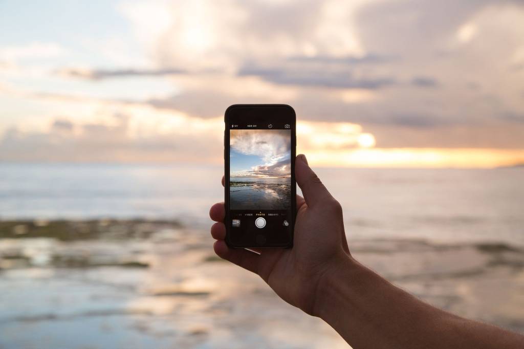 A imagem mostra a mão de uma pessoa segurando um Iphone, com uma praia ao fundo