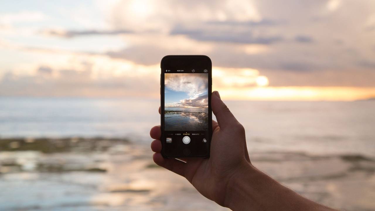 A imagem mostra a mão de uma pessoa segurando um Iphone, com uma praia ao fundo