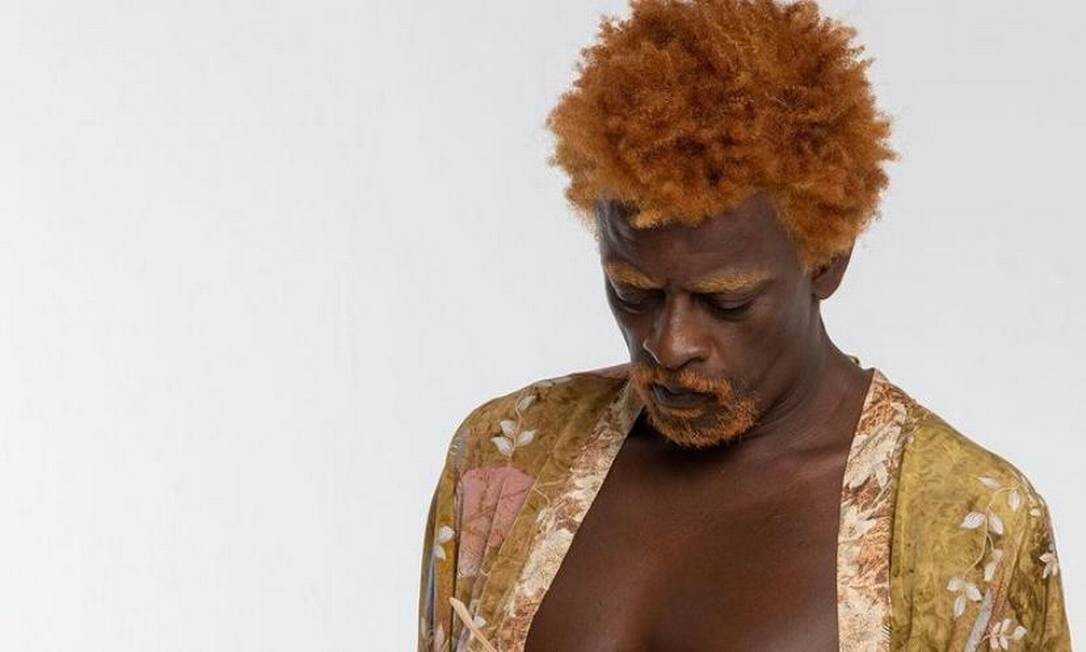 A imagem mostra o cantor Seu Jorge com os cabelos e a barba ruivos, com o peitoral à mostra, vestindo um quimono colorido, aberto na frente