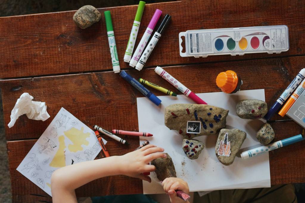 visão de uma mesa com tintas, canetas e uma criança pintando