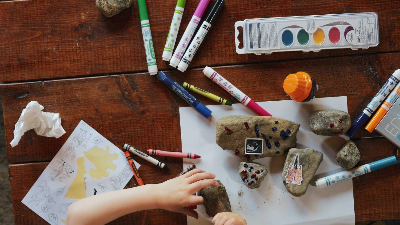 visão de uma mesa com tintas, canetas e uma criança pintando