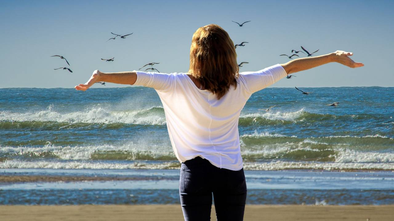 A imagem mostra uma mulher em frente ao mar com os braços abertos