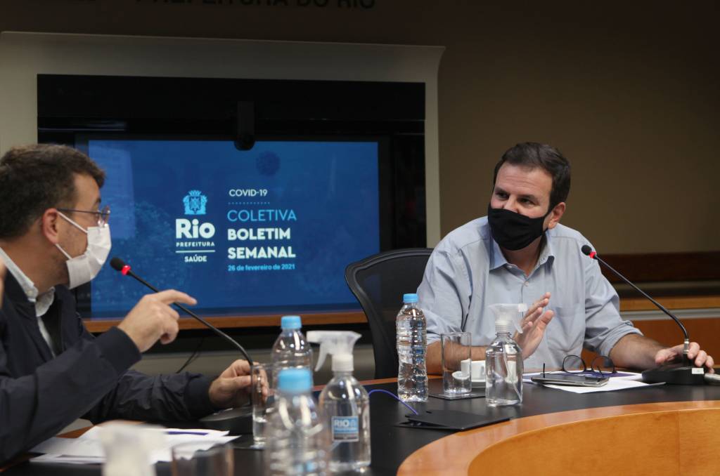 A imagem mostra o secretário de Saúde, Daniel Soranz, e o prefeito Eduardo Paes ao divulgar novo boletim epidemiológico, sentado à uma grande mesa de reunião