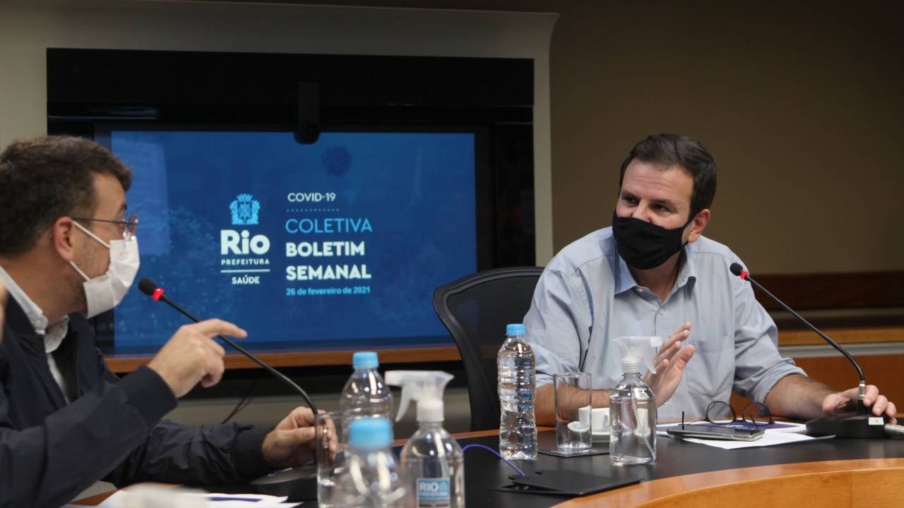 A imagem mostra o secretário de Saúde, Daniel Soranz, e o prefeito Eduardo Paes ao divulgar novo boletim epidemiológico, sentado à uma grande mesa de reunião