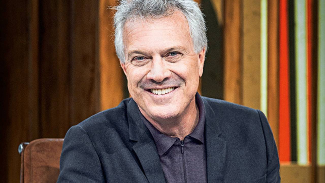 Homem com cabelo grisalho usando terno preto e camisa escura sorri
