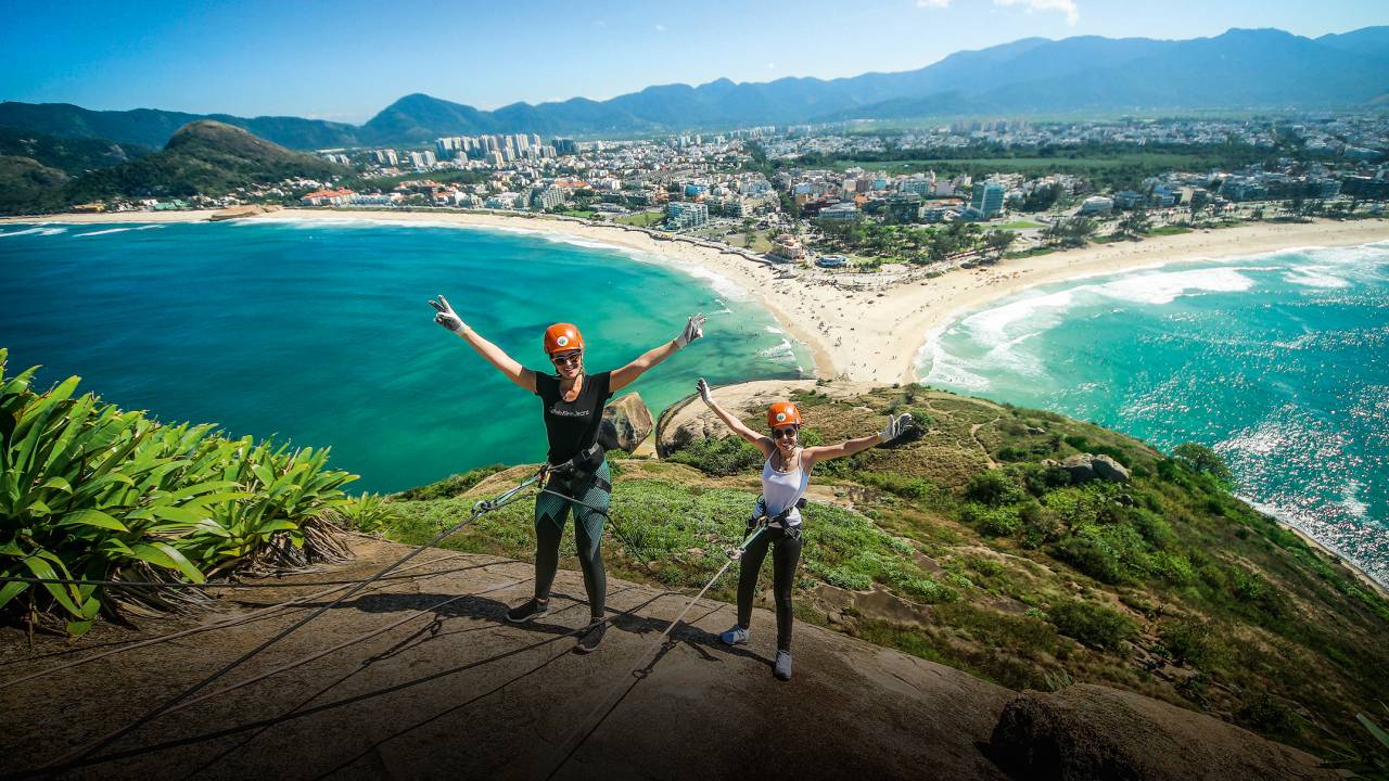 A imagem mostra duas moças descendo a Pedra do Pontal de rapel, com o mar embaixo