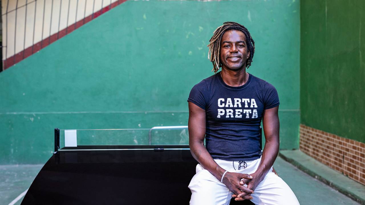 Homem negro com dreads usa camisa escura com os dizeres Carta Preta e calça branca. Ele está sentado e sorri