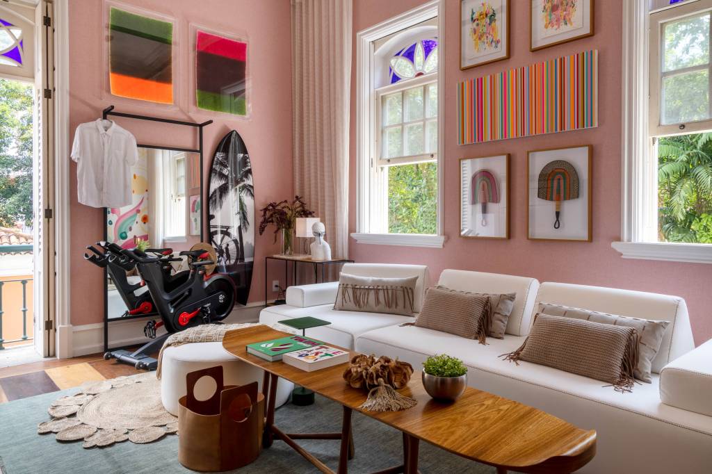 Saleta de estar em tons de rosa, quadros coloridos e uma bicicleta ergométrica