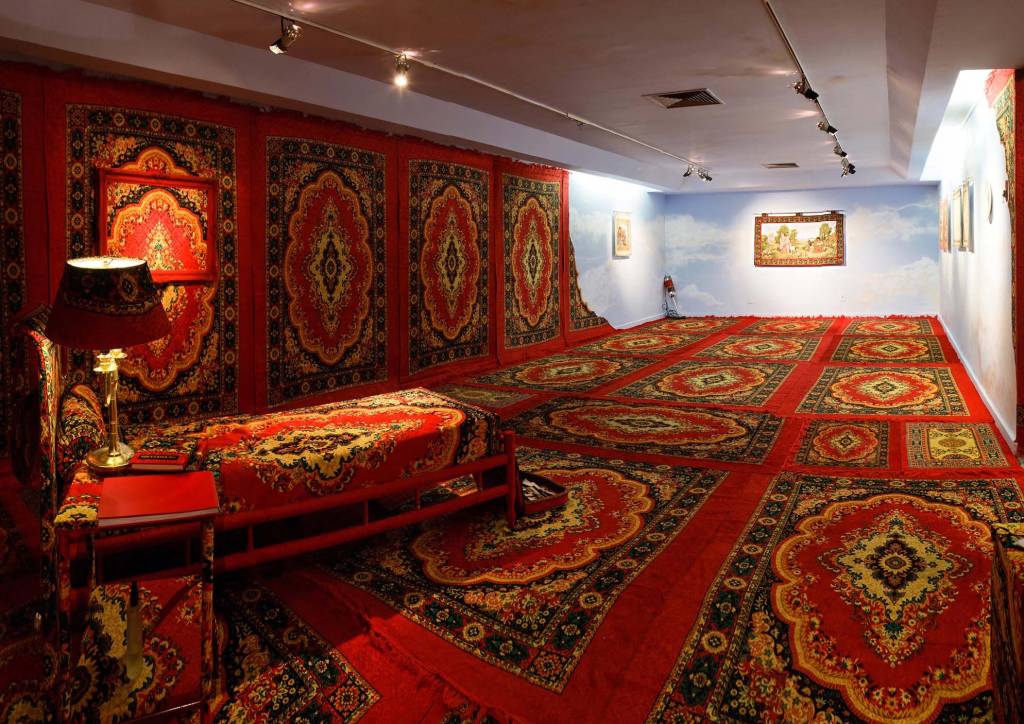 Claudia Hersz Sala da exposição na Casa França-Brasil com diversos tapetes persas pelas paredes e pelo chão