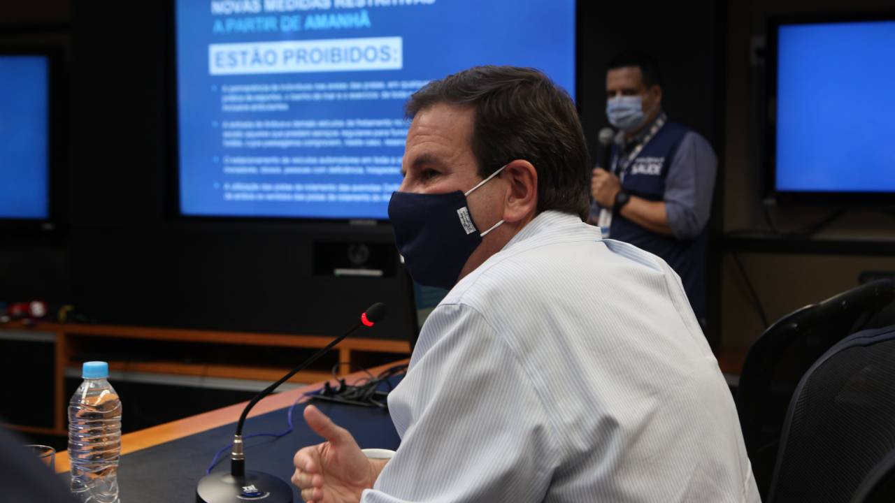 A imagem mostra o prefeito Eduardo Paes, de máscara, sentado à mesa, com um microfone à sua frente