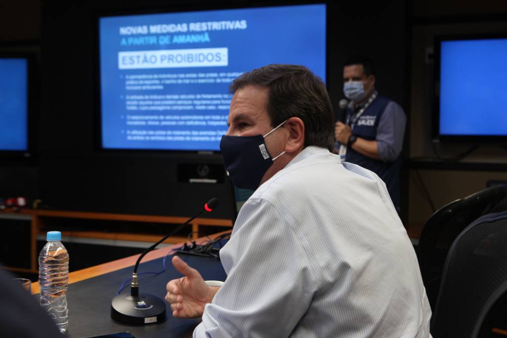 A imagem mostra o prefeito Eduardo Paes, de máscara, sentado à mesa, com um microfone à sua frente