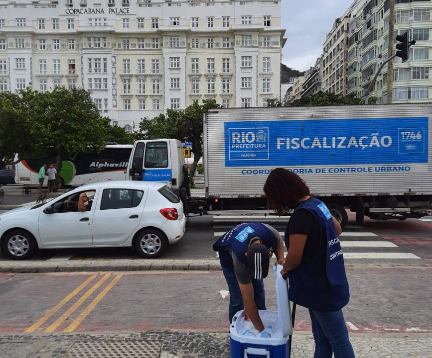 A imagem mostra uma fiscal dda Vigilância Sanitária abordando um vendedor de cerveja no calçadão, em frente ao Copacabana Palace
