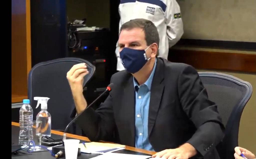 A imagem mostra o prefeito Eduardo Paes dando entrevista, sentado, usando máscara