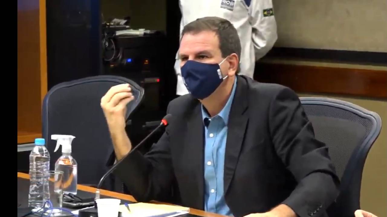 A imagem mostra o prefeito Eduardo Paes dando entrevista, sentado, usando máscara