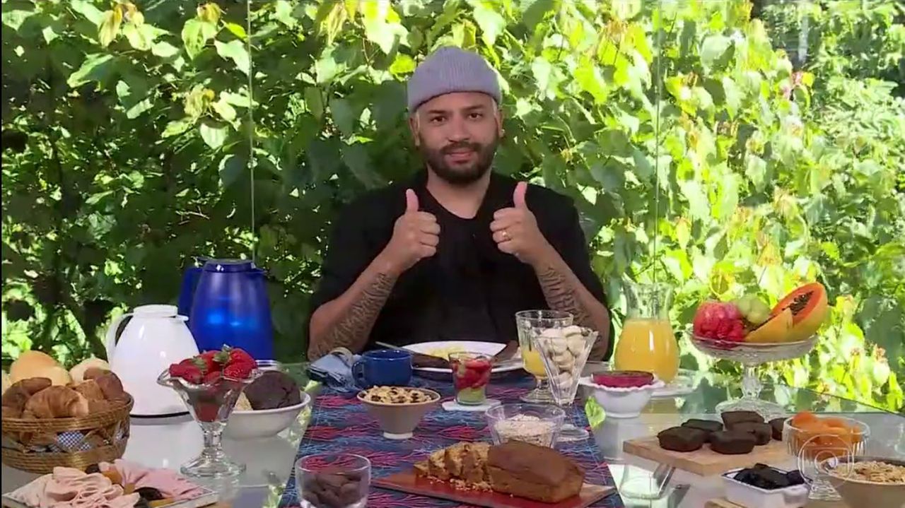 A imagem mostra o rapper Projota fazendo sinal de positivo, à mesa do café da manhã de Ana Maria Braga