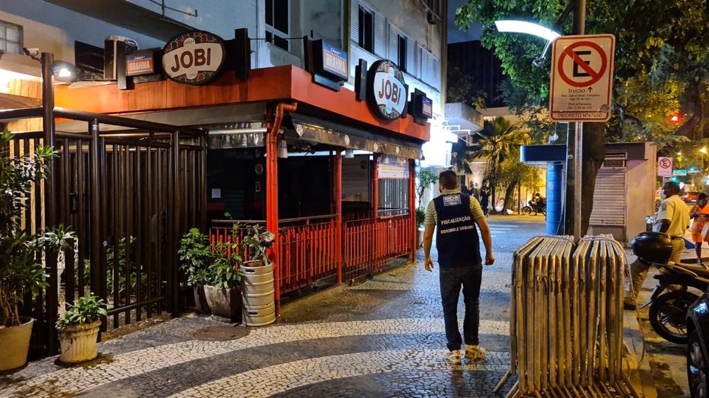 A imagem mostra um fiscal da prefeitura passando em frente ao bar Jobi, no Leblon, que está com as portas fechadas