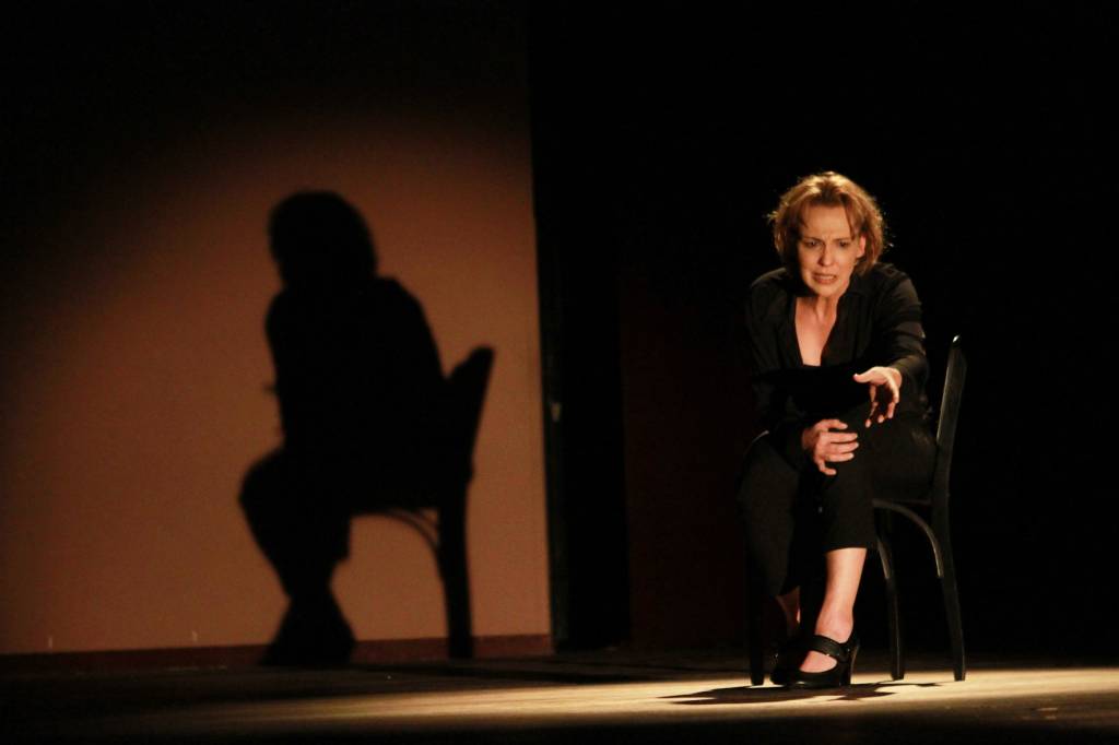 Ana Beatriz Nogueira em cena, sentada numa cadeira, com sua sombra projetada ao lado