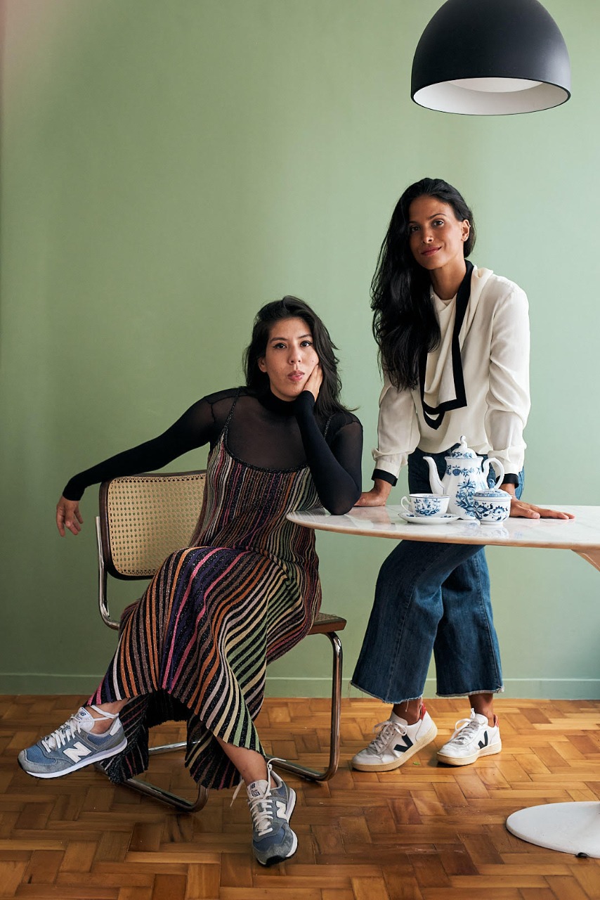 Duas mulheres brancas com cabelos longos castanhos posam na foto, uma em pé com blusa branca, outra sentada com blusa preta