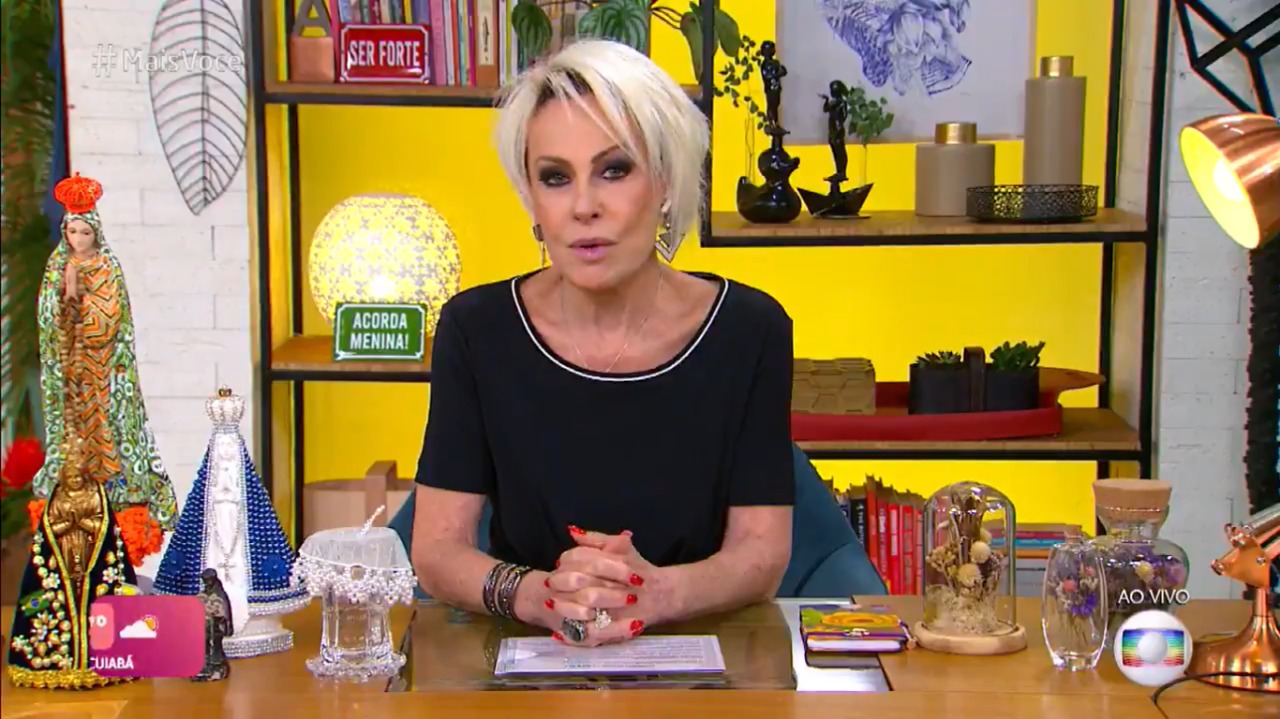 A imagem mostra a apresentadora Ana Maria Braga na bancada de seu programa, com uma parede amarela ao fundo e as mãos cruzadas sobre a mesa
