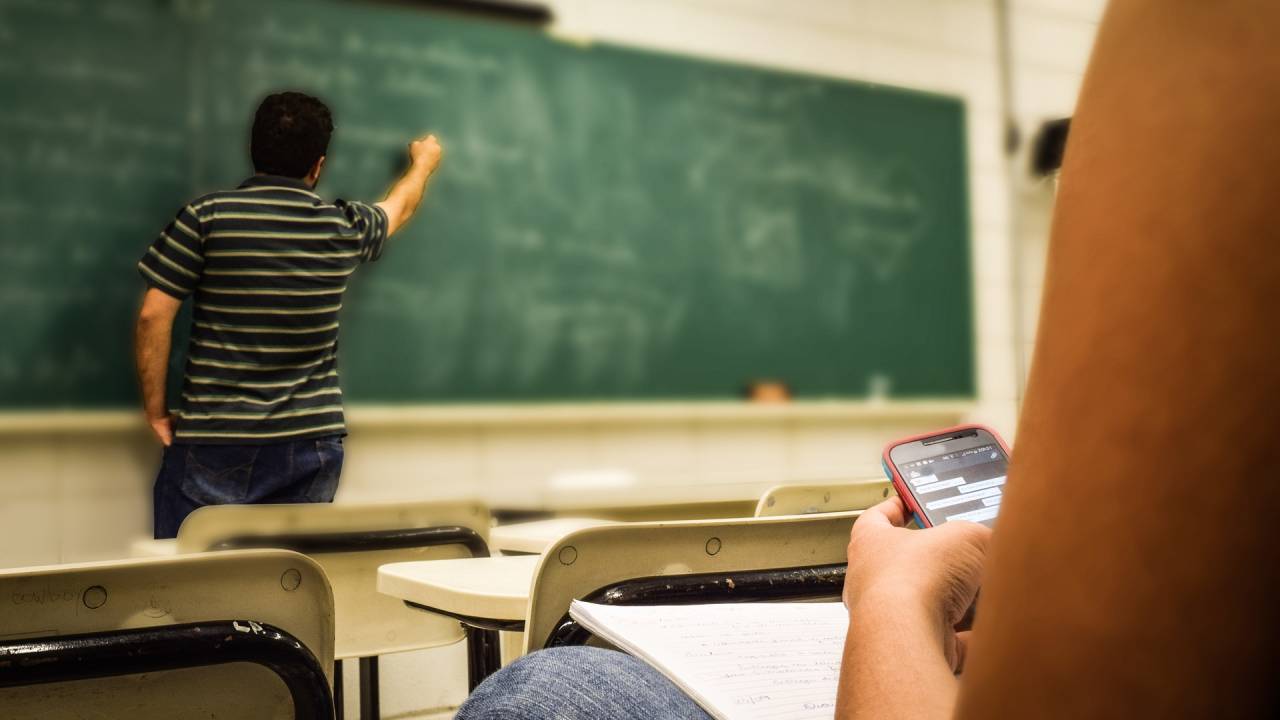 A imagem mostra um professor escrevendo no quadro e uma aluna sentada