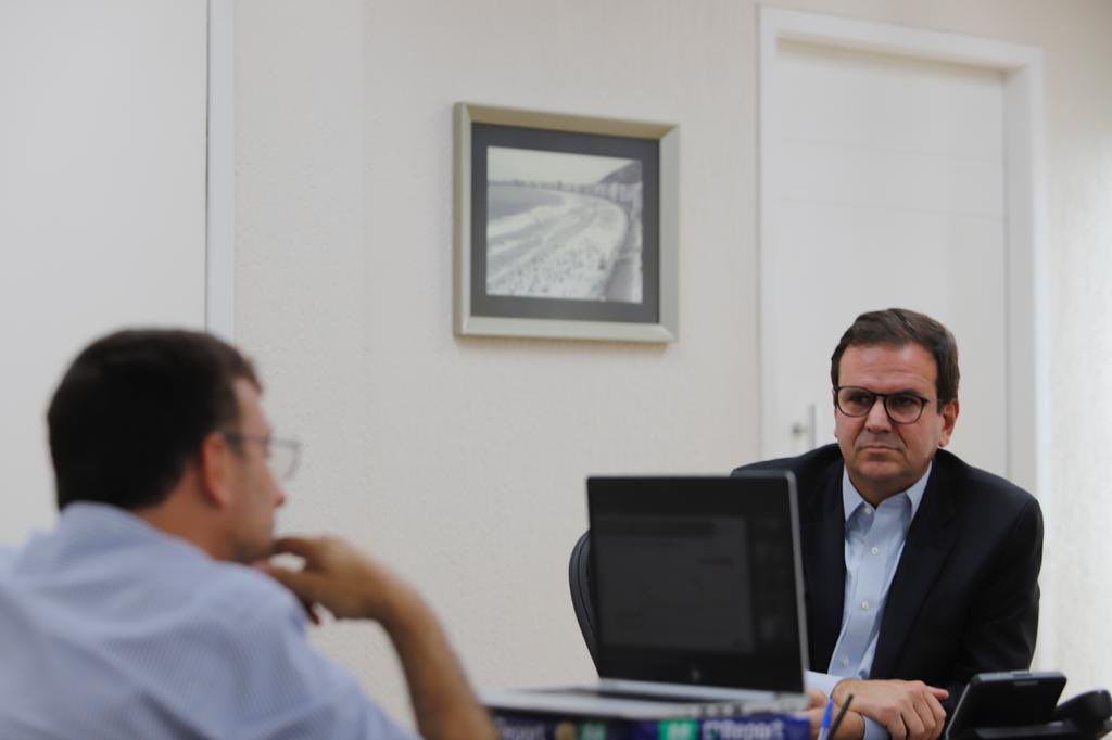 A imagem mostra o prefeito Eduardo Paes sentado, de frente para a câmera com o secretário de saúde Daniel Soranz, de costas