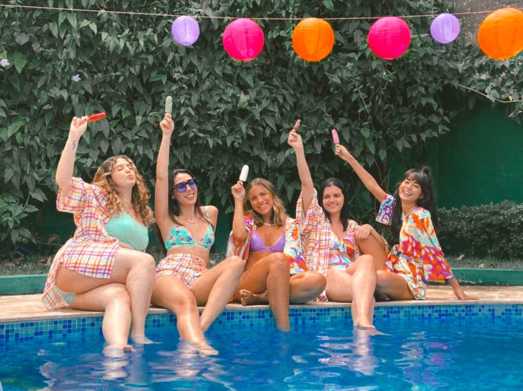 Cinco meninas usando biquini estão sentadas à beira da piscina