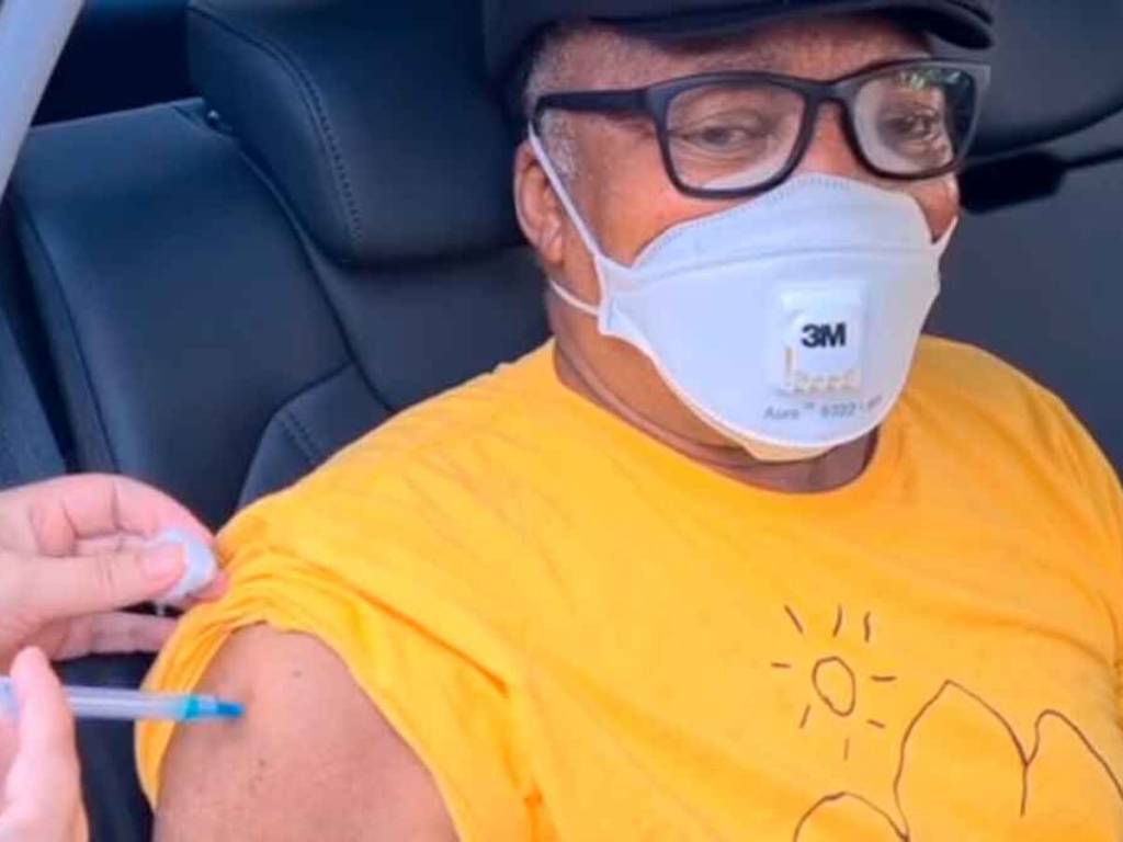 A imagem mostra Milton Nascimento, de óculos, boné, e camiseta amarela, sendo vacinado dentro do carro