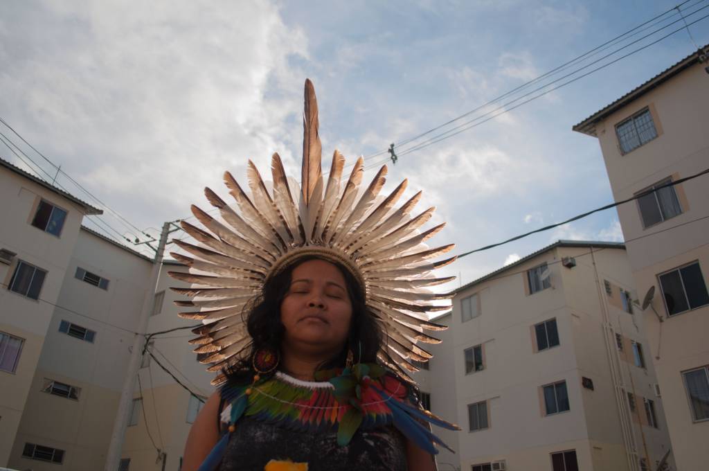 Mulher indígena com cocar sob o sol. Atrás dela, um prédio