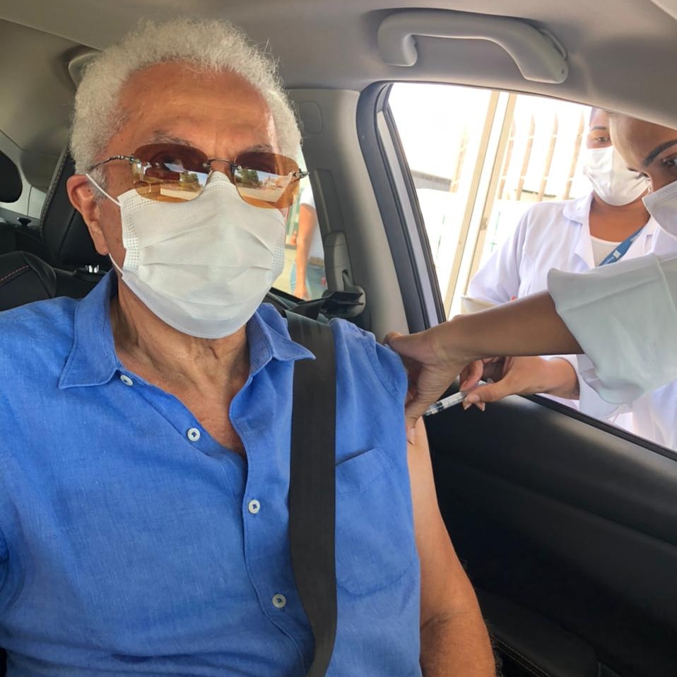 A imagem mostra Paulinho da Viola, de camisa azul, sendo vacinado dentro do carro