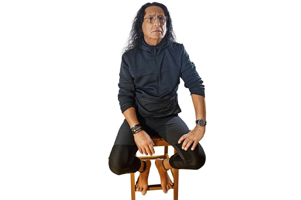 Gerald Thomas, de cabelos compridos, sentado em um banquinho de madeira