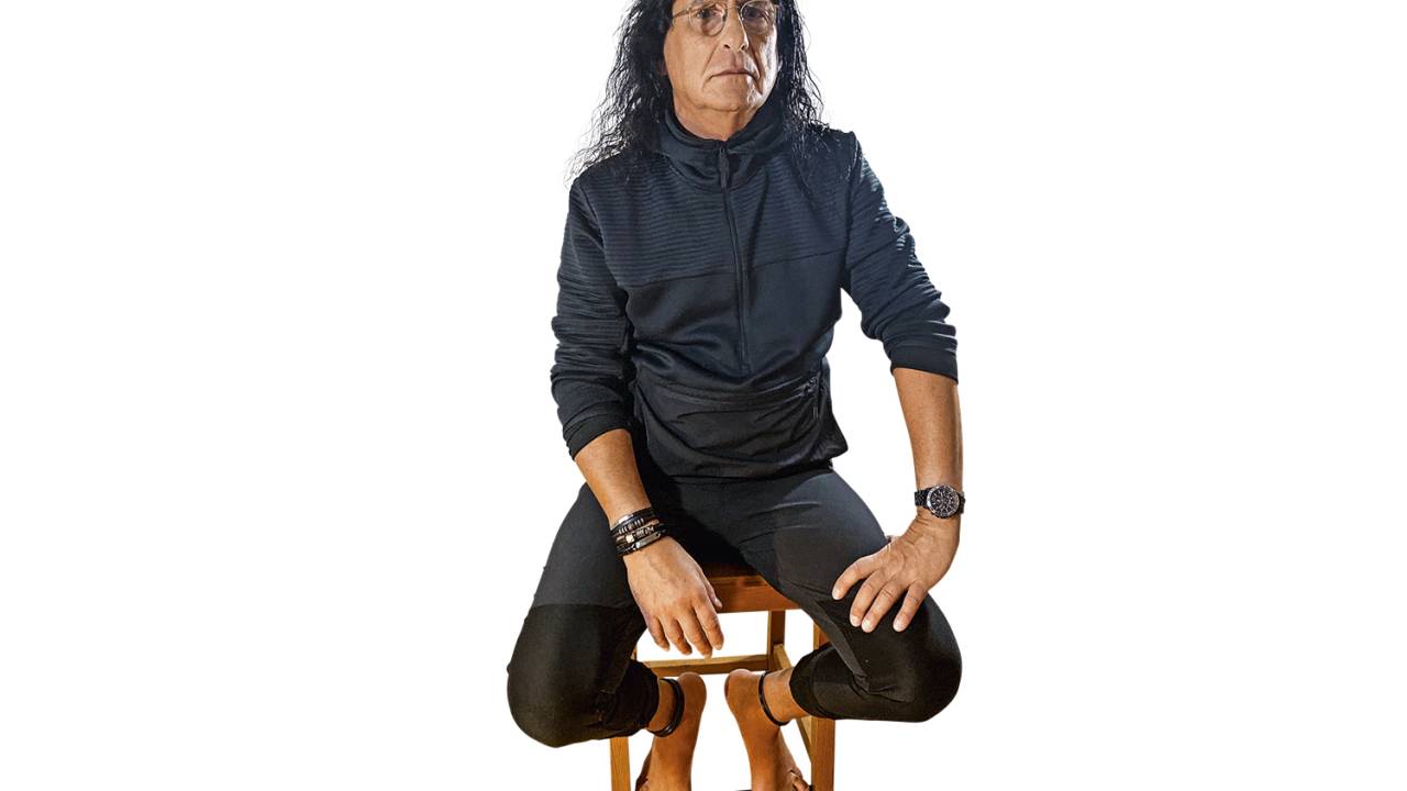 Gerald Thomas, de cabelos compridos, sentado em um banquinho de madeira