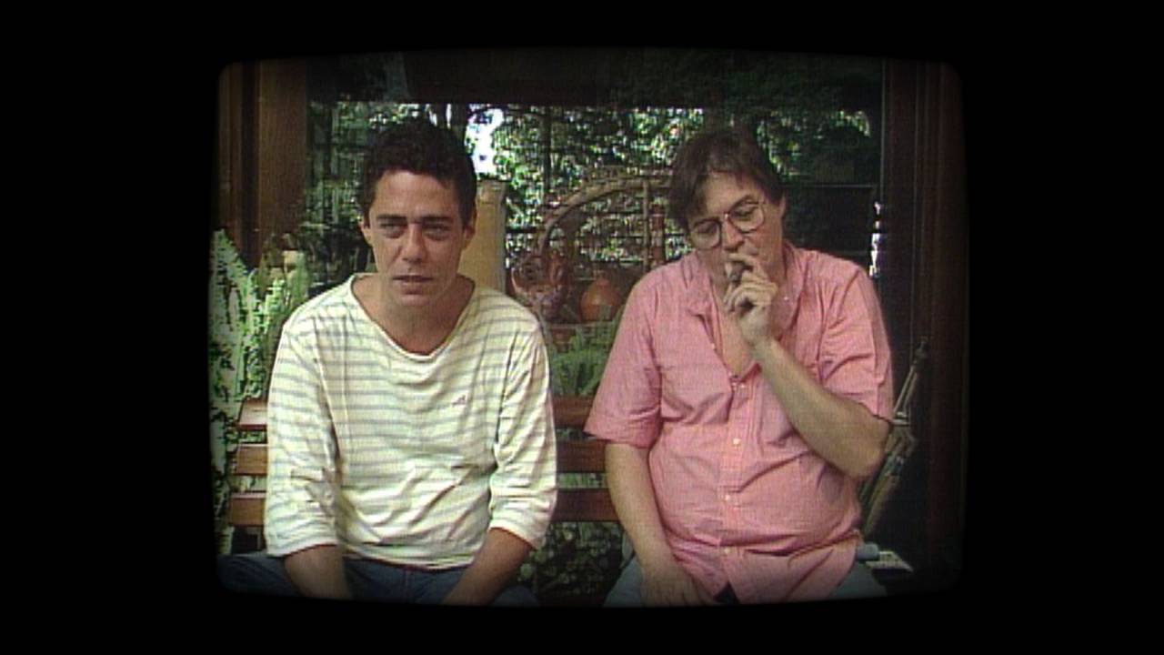 A imagem mostra Chico Buarque e Tom Jobim, sentados lado a lado num jardim, nos final dos anos 80; Tom fuma charuto